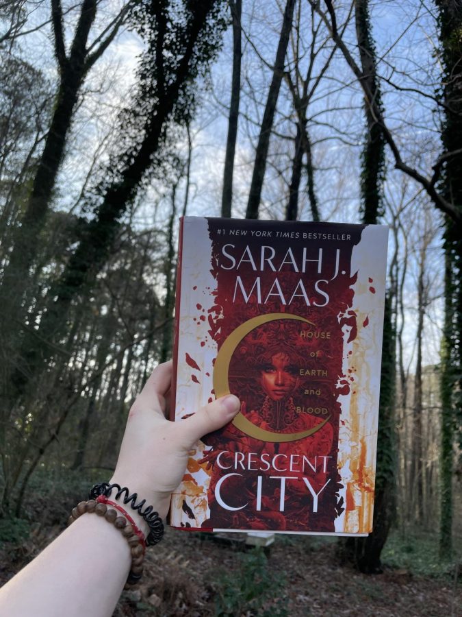 A hand holding House of Earth and Blood, Sarah J. Mass, in the woods.