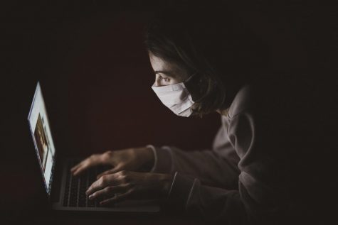 A student in a dark room wearing a covid-19 mask, staring at her computer screen.
