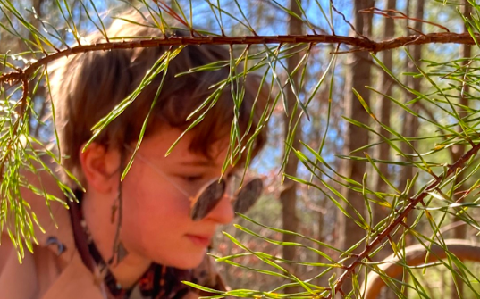 Raine Tillotson looks towards a plant off-camera. They are in a forest, surrounded by tree branches. (Photo courtesy of Raine Tillotson)