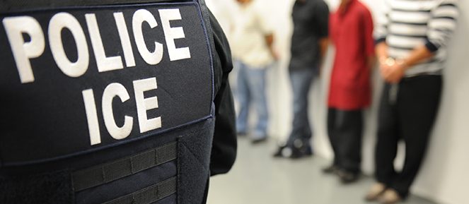 A closeup image of an ICE agent’s vest, reading “POLICE ICE.” Several people in average clothing are lined up against a wall in the background. (Photo is in the public domain, courtesy of ICE)