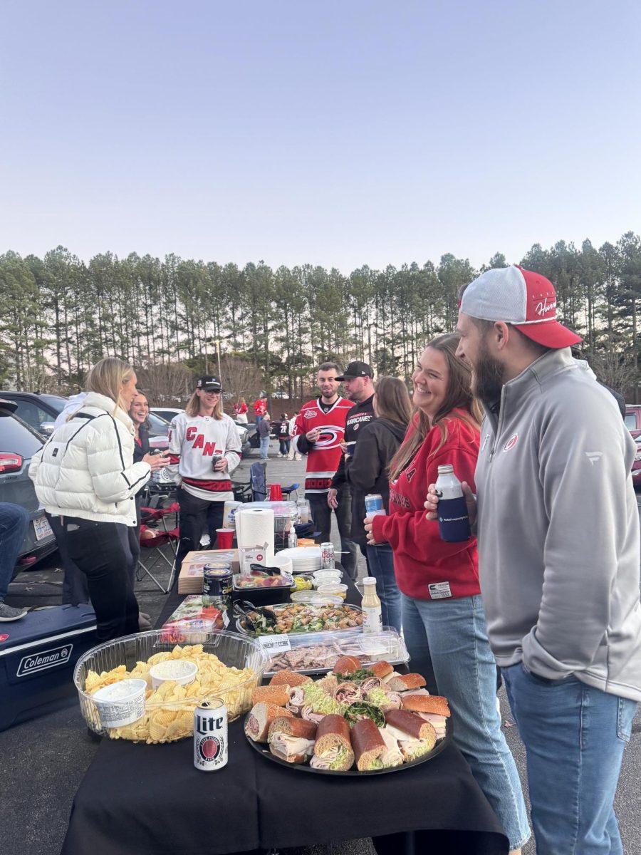 A group of Hurricanes fans are seen conversing around a table with various dishes on it. Many Hurricanes fans take their tailgates to the next level by bringing delectables to their tailgates.