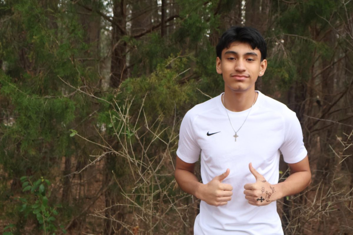 Christian Dominguez poses for a picture, wearing an athletic shirt. He was stood in front of a group of trees next to the main Athens Drive High School building.