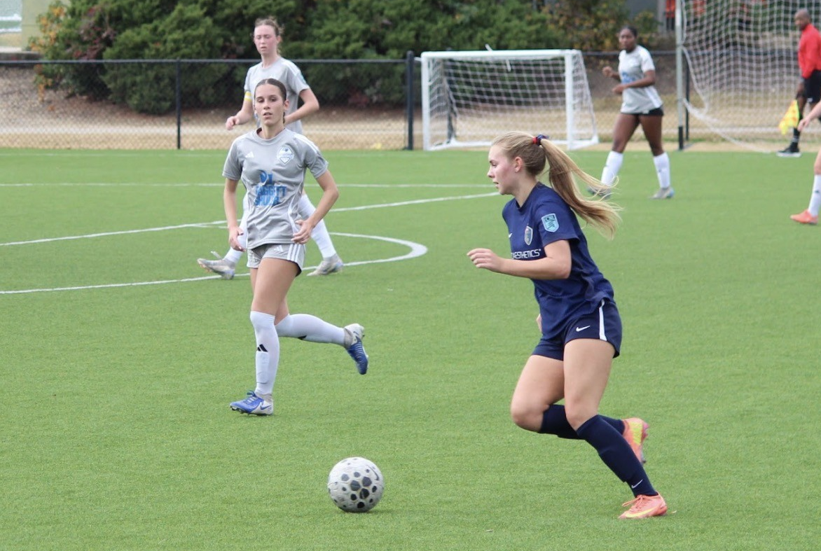Gracie De Haro during a soccer game, with possession of the ball. She looks out across the field, prepared to make her next move as the opposing team closes in. 