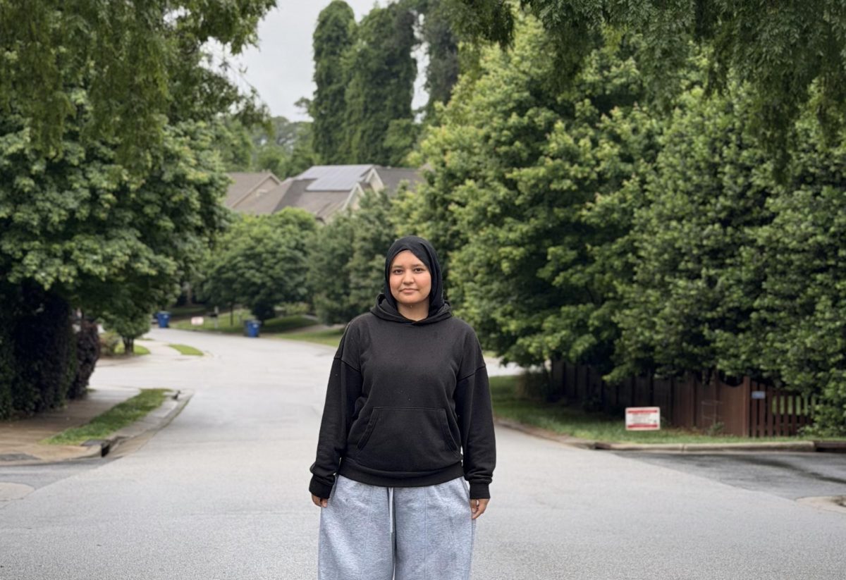 A quiet moment of stillness finds Hania standing at the center of a lush, suburban landscape. Framed by towering greenery and soft morning light, she carries an air of calm confidence against the backdrop of the neighborhood.
